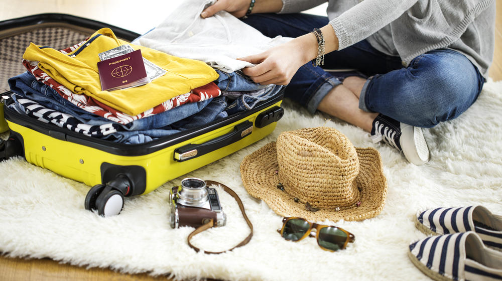 Frau sitzt am Boden auf einem Teppich, vor sich ein gelber Koffer. Darin befinden sich schon Kleidungsstücke, ein Reisepass. Sonnenhut, Strandschuhe, Sonnenbrille liegen noch auf dem Teppich.