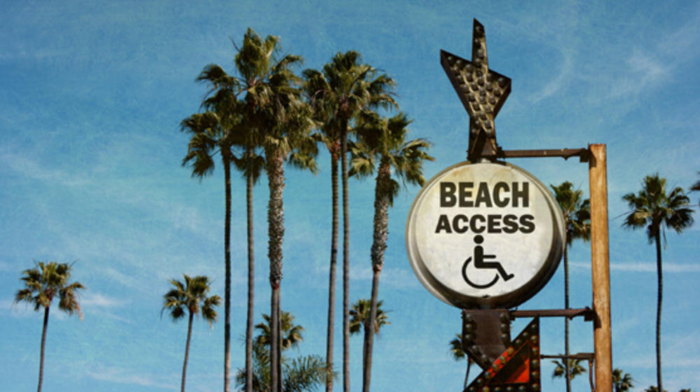 Hohe Palmbäume vor blauem Himmel, davor ein Schild mit einem gezeichneten Rollstuhl und Schrift: Beach Access.