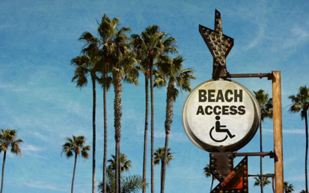 Hohe Palmbäume vor blauem Himmel, davor ein Schild mit einem gezeichneten Rollstuhl und Schrift: Beach Access.