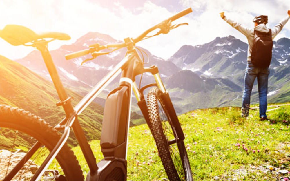 In den Bergen ein Fahrrad im Vordergrund, dahinter ein Mann, die Arme weit von sich gestreckt, die Sonne scheint.