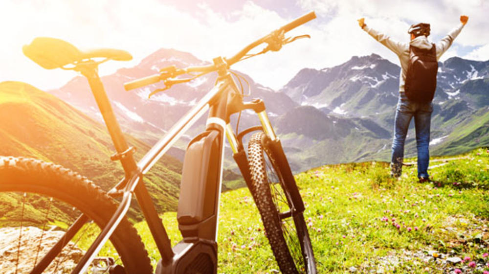 In den Bergen ein Fahrrad im Vordergrund, dahinter ein Mann, die Arme weit von sich gestreckt, die Sonne scheint.