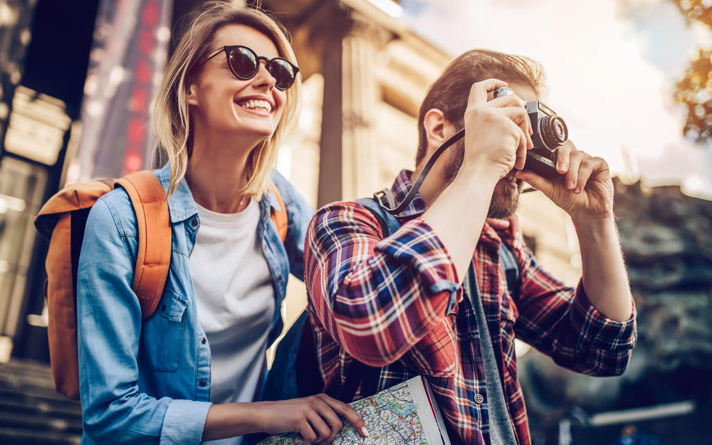 Touristenpärchen, sie mit Sonnenbrille lächelnd, er hält einen Fotoapparat zum Knipsen bereit.