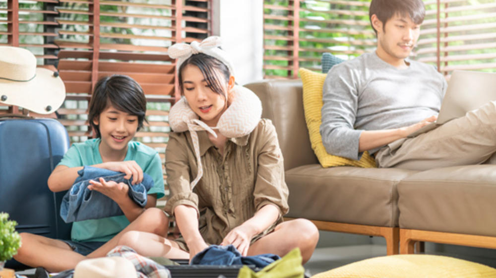 Mutter und Sohn sitzen am Boden und packen einen Koffer, der Vater sitzt am Sofa am Laptop.
