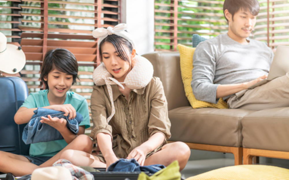 Mutter und Sohn sitzen am Boden und packen einen Koffer, der Vater sitzt am Sofa am Laptop.
