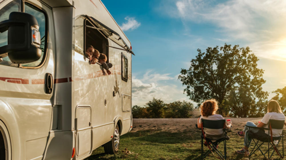 Rückansicht eines jungen Paares auf Klappsesseln neben einem Campingbus, aus einem der Fenster schauen drei kleine Kinder.