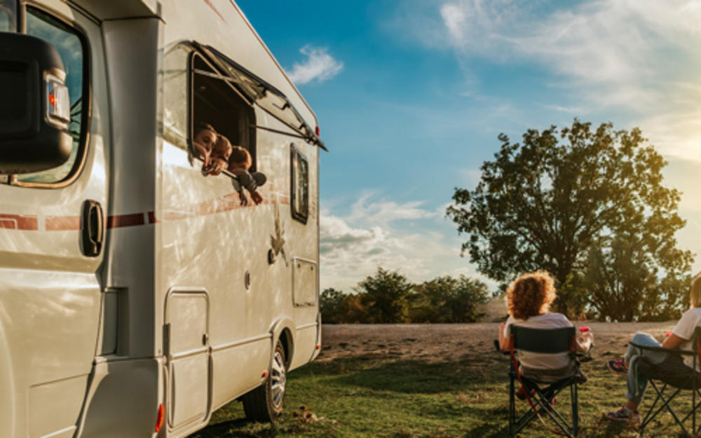 Rückansicht eines jungen Paares auf Klappsesseln neben einem Campingbus, aus einem der Fenster schauen drei kleine Kinder.