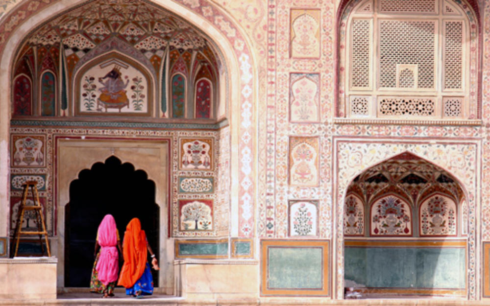 Frontansicht eines arabischen Bauwerks welches künstlerisch bemalt und verziert ist, in einem Tor die Rückansicht zweier bunt gekleideter Frauen.