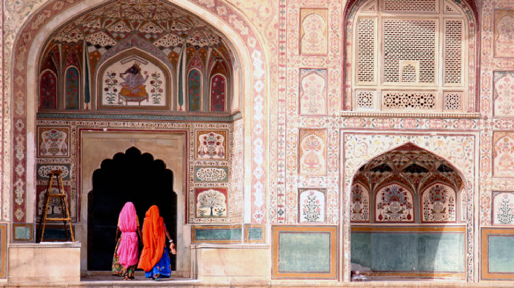 Frontansicht eines arabischen Bauwerks welches künstlerisch bemalt und verziert ist, in einem Tor die Rückansicht zweier bunt gekleideter Frauen.