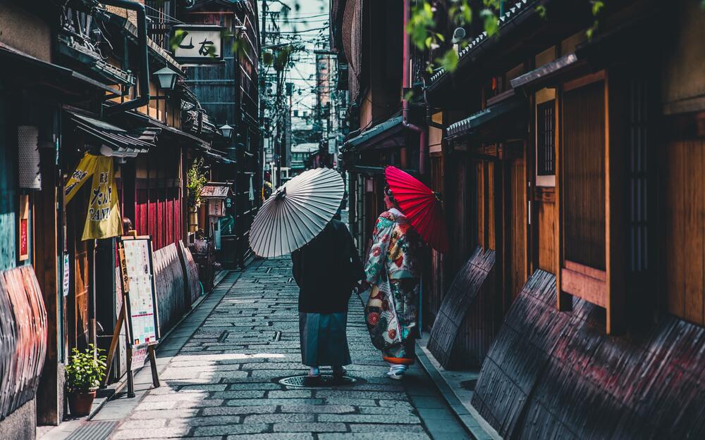 Two ladies in traditional Japanese attire and with umbrellas are walking on the streets of Kyoto.