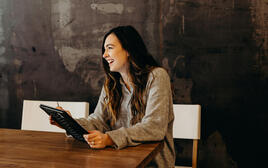A woman sits relaxed at a table, holding a tablet in her hands.