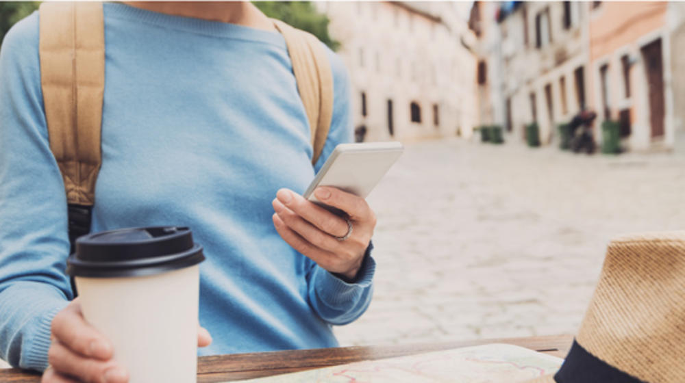 Woman in light blue sweater with backpack holds a coffee cup in one hand, a cell phone in the other.