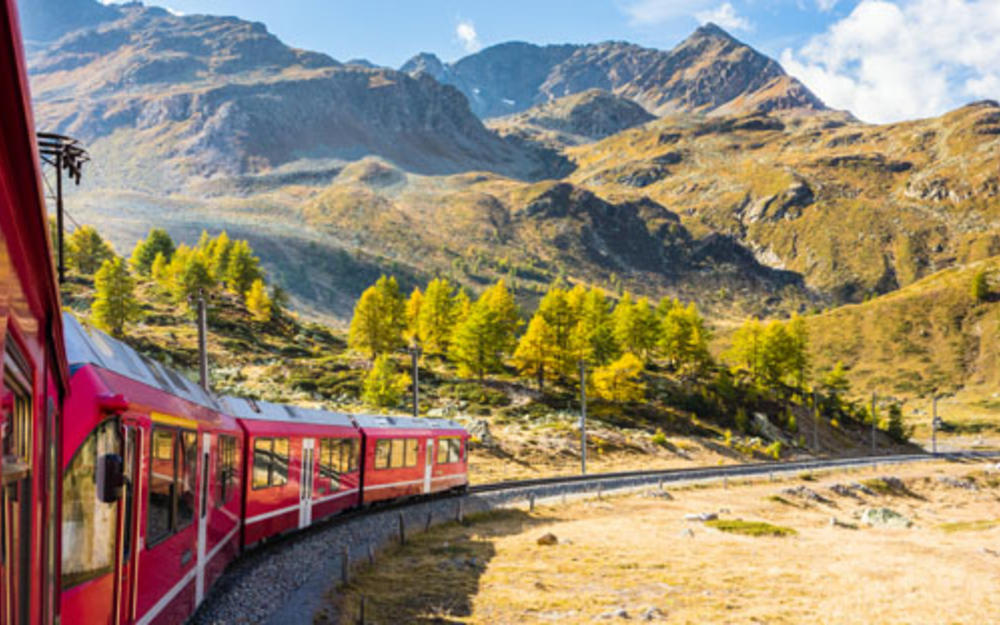 Auf der linken Bildhälfte ist ein langer, roter Zug zu sehen, vor einer hochgelegenen Berglandschaft.