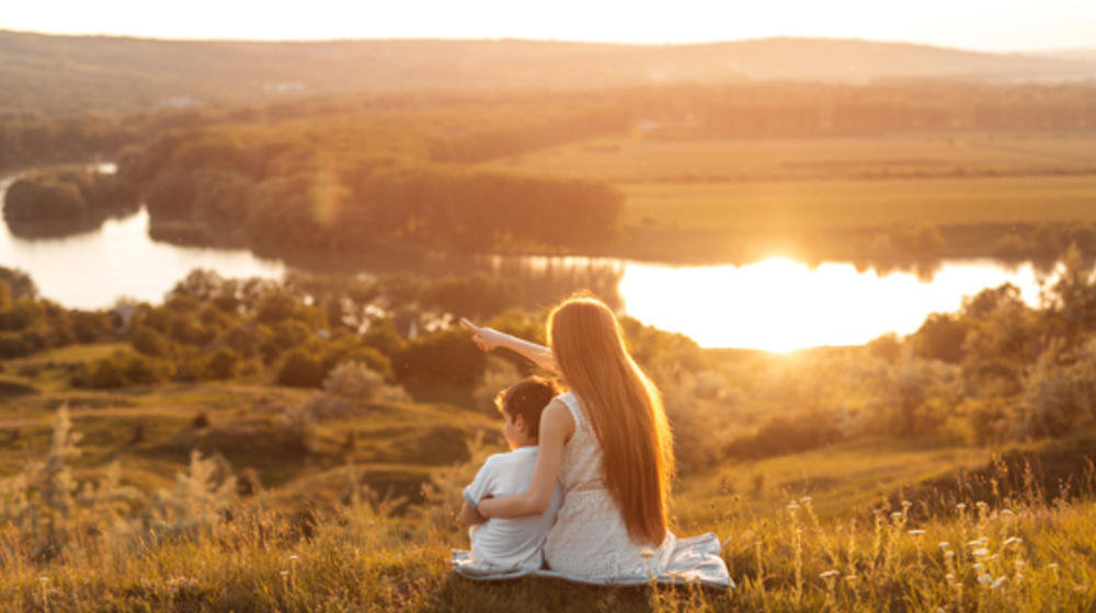 Rückansicht einer jungen Frau mit kleinem Jungen bei der Betrachtung des Sonnenuntergangs mit ruhigem Fluß und wogende Felder.