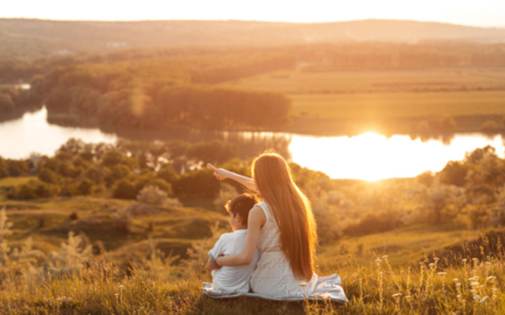 Rückansicht einer jungen Frau mit kleinem Jungen bei der Betrachtung des Sonnenuntergangs mit ruhigem Fluß und wogende Felder.