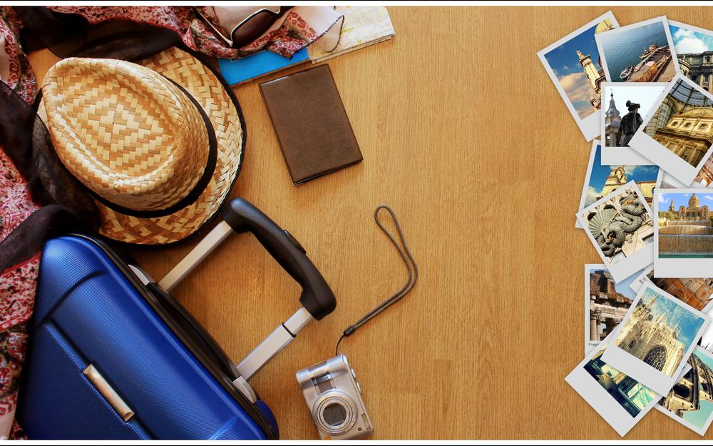 Am Boden liegen diverse Fotos von Reisezielen, ein Strohhut, daneben ein Koffer.