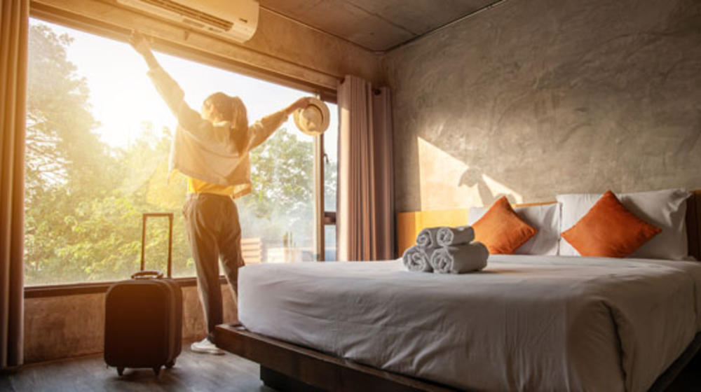 Junge Frau genießt die Abendsonne bei der Ankunft in ihrem Hotelzimmer und streckt dabei die Arme aus.