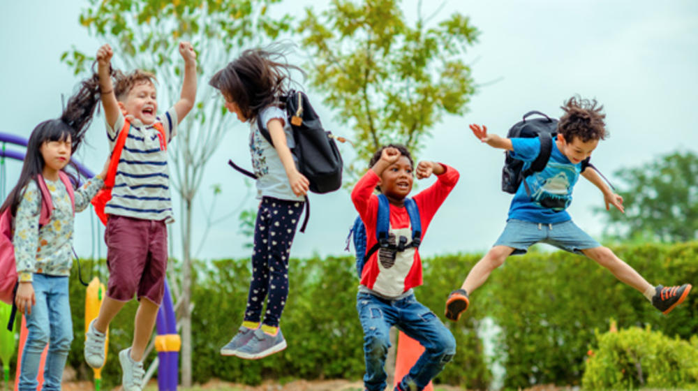 Fünf Erstklassler mit Schultasche am Rücken springen vor Freude in die Luft.