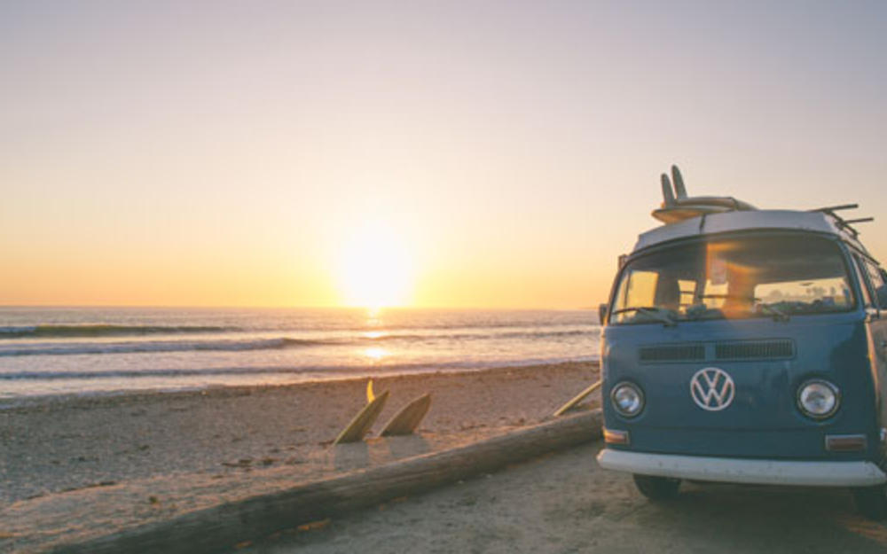Ein hellblauer VW Van parkt am Strand bei Sonnenuntergang.