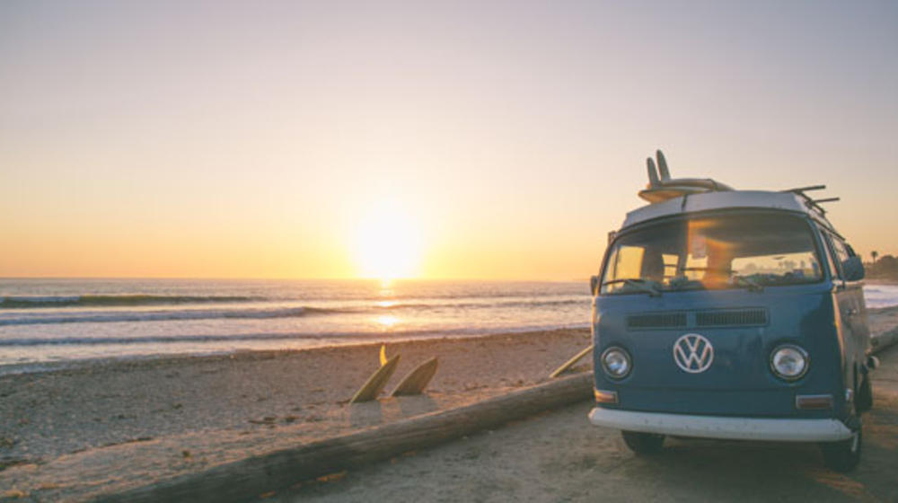 Ein hellblauer VW Van parkt am Strand bei Sonnenuntergang.