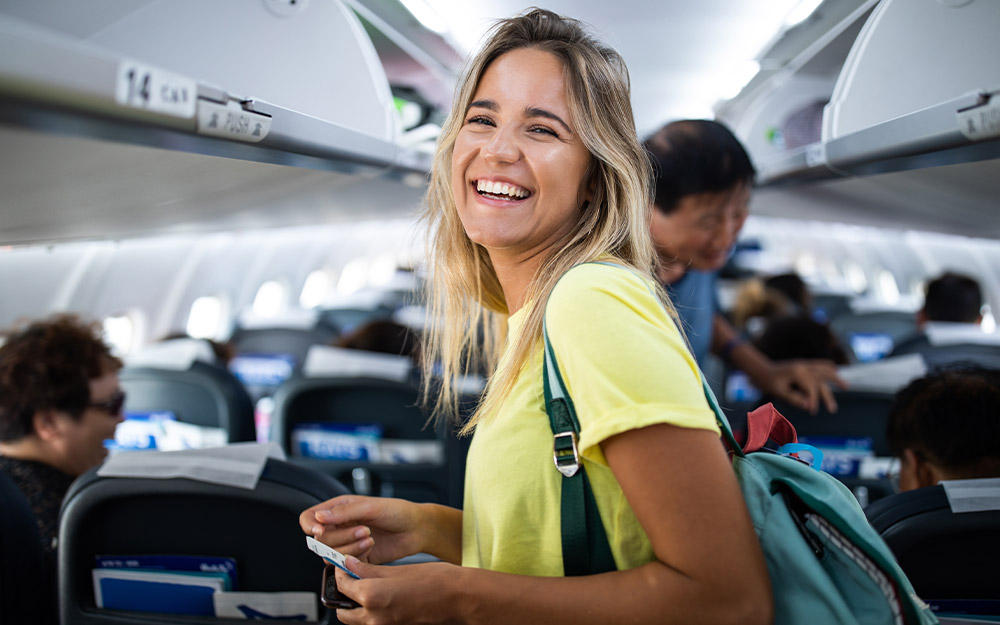 Junge lachende Frau steht im Gang eines Fliegers und sucht ihren Sitzplatz