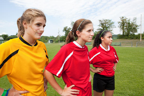Drei junge Fußballspielerinnen am Rasen, zwei davon in Trikots rot-weiß, eine im Trikot gelb-schwarz, Hände in die Hüften gestemmt.