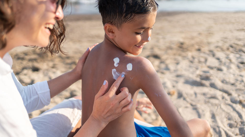 The mother is applying sunscreen to her little son on the beach.