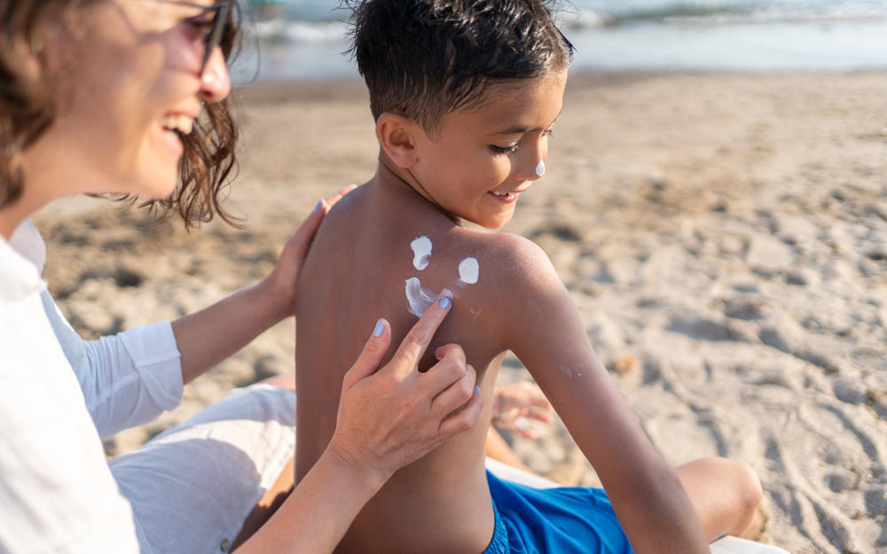 The mother is applying sunscreen to her little son on the beach.