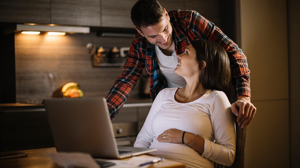 Young expectant parents sit in front of a laptop and plan their babymoon
