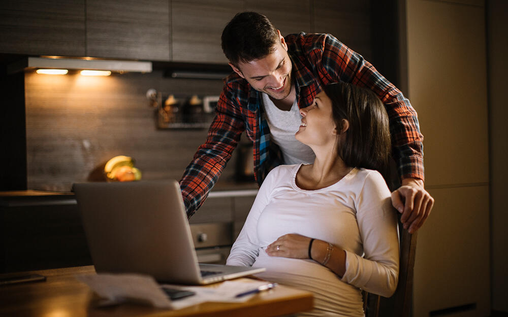 Young expectant parents sit in front of a laptop and plan their babymoon