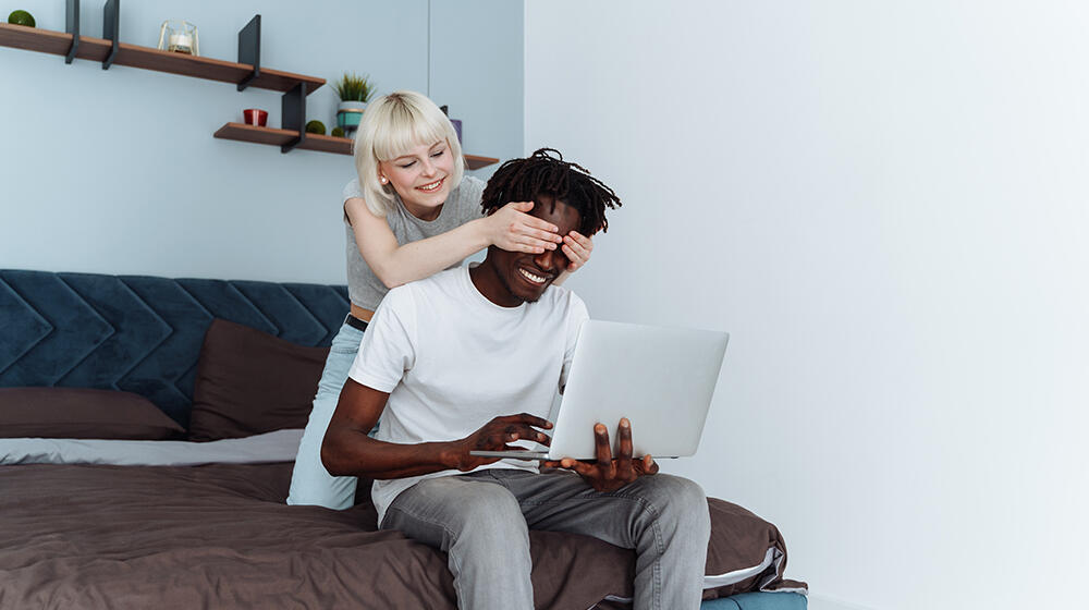 Young woman surprises her smiling boyfriend and covers his eyes while he is typing on his laptop