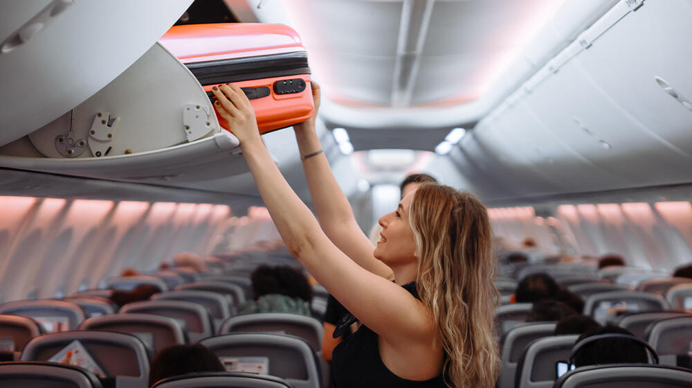 A woman stows her carry-on suitcase in the airplane’s overhead compartment