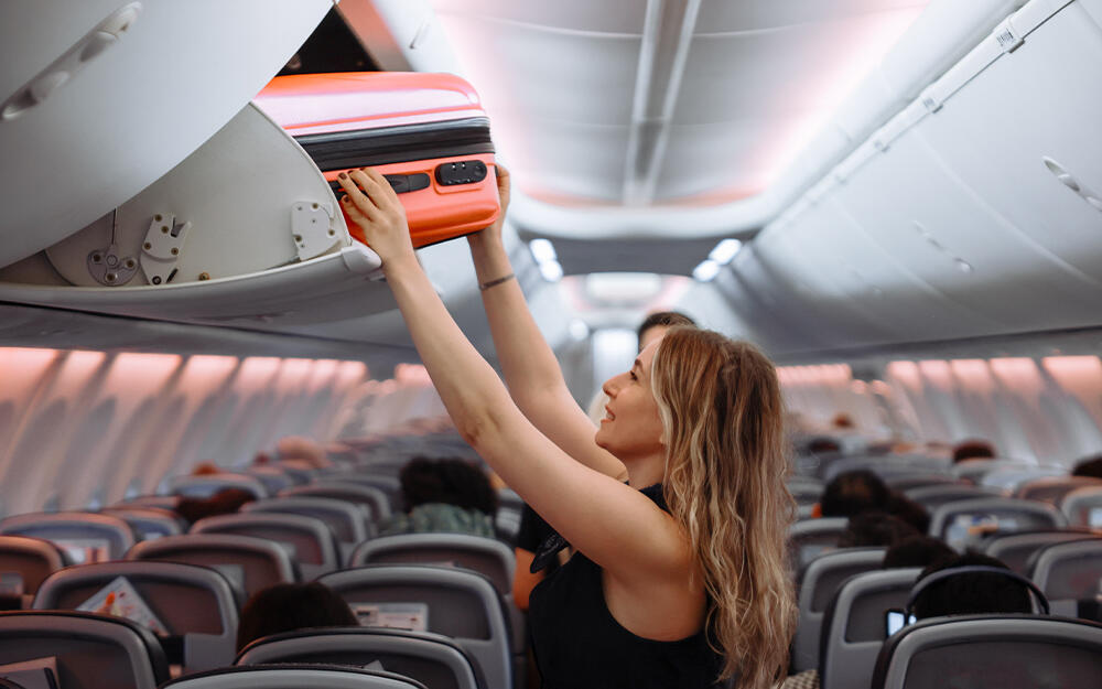 A woman stows her carry-on suitcase in the airplane’s overhead compartment