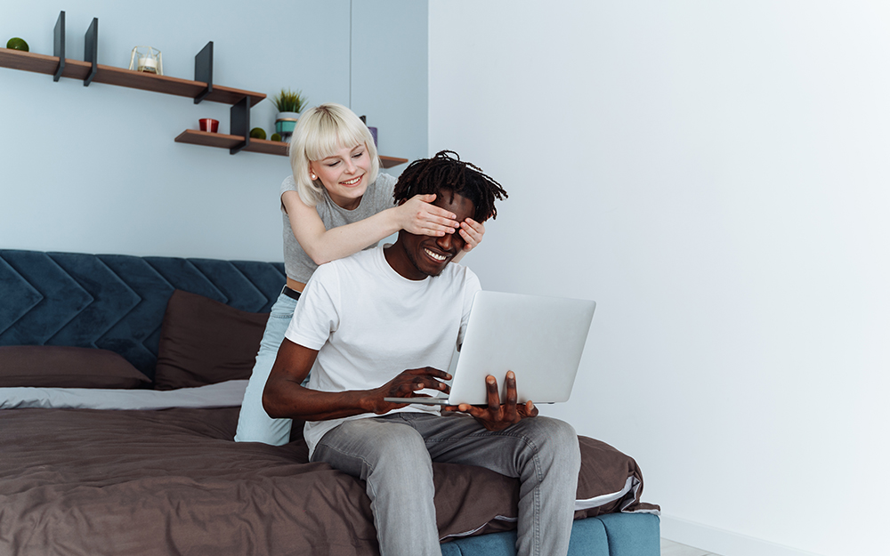 Young woman surprises her smiling boyfriend and covers his eyes while he is typing on his laptop